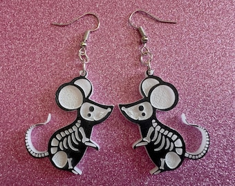 May include: A pair of silver skeleton mouse earrings. The mice are black with white bones and white ears. The earrings are on a pink sparkly background.