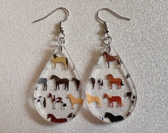 May include: Clear, teardrop-shaped earrings with a variety of horse illustrations. The earrings have silver-colored hooks and feature multiple horse breeds in various colors, including brown, black, and tan. The earrings are lightweight and suitable for everyday wear.