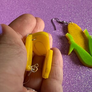 May include: A pair of yellow corn on the cob earrings. One earring is shown in detail, with the back of the earring visible. The other earring is shown from the front.