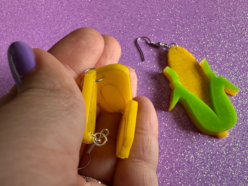 May include: A pair of yellow corn on the cob earrings. One earring is shown in detail, with the back of the earring visible. The other earring is shown from the front.