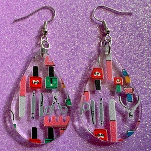 May include: Clear, teardrop-shaped earrings with a nail salon theme. The earrings feature a pattern of nail polish bottles, nail files, scissors, and other manicure tools in red, green, pink, and gray. The earrings have silver-colored hooks.