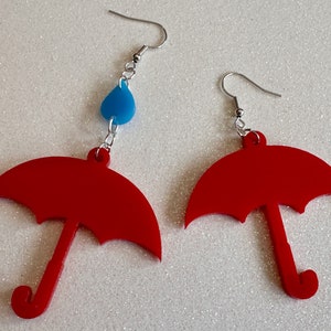 May include: A pair of red umbrella earrings with a blue raindrop charm on one earring.