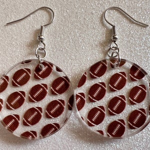 May include: A pair of clear acrylic earrings with a brown and white football pattern. The earrings have silver hooks.