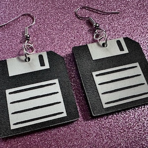 May include: A pair of black floppy disk earrings with silver-colored accents. Each earring features a square shape with a rectangular top and horizontal lines, suspended from silver-tone ear wires.