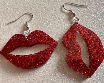 May include: A pair of red glitter earrings shaped like lips. One earring is a solid lip shape, while the other is a more abstract lip shape with a cutout.