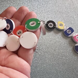 May include: A set of 12 white, round, felt earrings with black numbers 1 through 12. The earrings are in the shape of billiard balls and are strung together on a silver chain.