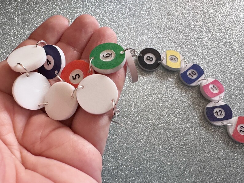 May include: A set of 12 white, round, felt earrings with black numbers 1 through 12. The earrings are in the shape of billiard balls and are strung together on a silver chain.
