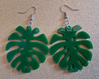 May include: A pair of green monstera leaf earrings with silver hooks. The leaves are cut out and have a glossy finish.