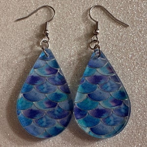Puede incluir: Un par de pendientes en forma de lágrima con un diseño de escamas de sirena. Los pendientes presentan un patrón de escamas azules, turquesas y moradas. Los pendientes tienen ganchos plateados.