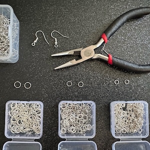 Puede incluir: Un conjunto de suministros para hacer joyas que incluye ganchos para pendientes, anillos de salto y alicates en un recipiente de plástico transparente sobre una superficie negra.