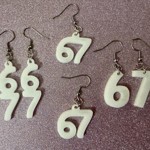 Puede incluir: Pendientes blancos con el número 67 y ganchos plateados. Los pendientes son de estilo sencillo y minimalista. Se muestran sobre una superficie texturizada de color púrpura claro.