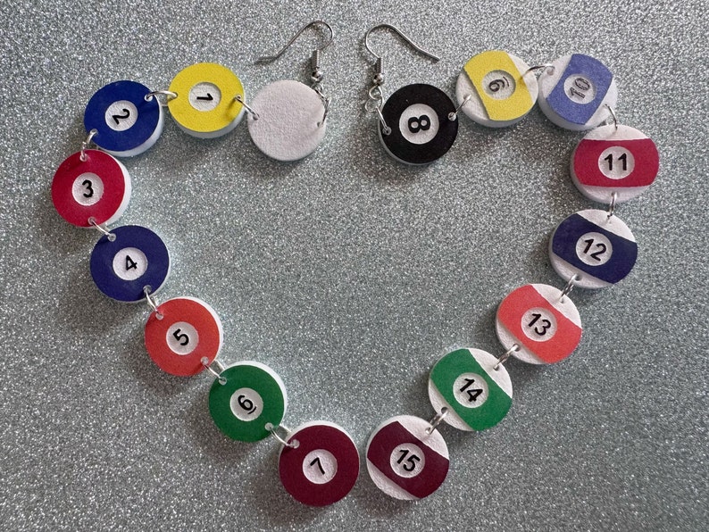 May include: A pair of earrings with white and black billiard ball charms. The charms are numbered 1 through 15 and are connected by silver jump rings. The earrings are hanging from silver hooks.