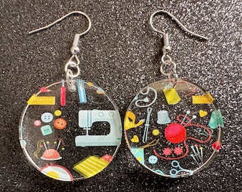 May include: A pair of silver earrings with clear resin circles. The circles are decorated with a colorful pattern of sewing-themed imagery, including a sewing machine, buttons, needles, and thread.