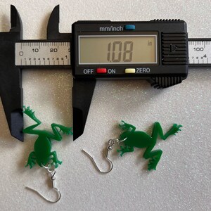 May include: A pair of green frog earrings with silver hooks. The earrings are measured with a caliper, showing a measurement of 1.08 inches.