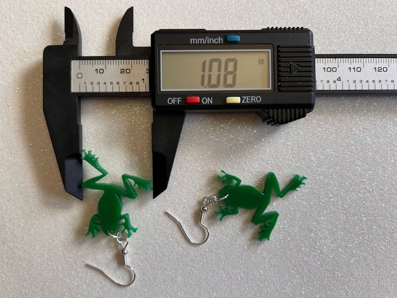 May include: A pair of green frog earrings with silver hooks. The earrings are measured with a caliper, showing a measurement of 1.08 inches.