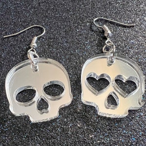 May include: A pair of silver skull earrings with heart-shaped cutouts for the eyes. The earrings are made of clear acrylic and have a mirror finish.
