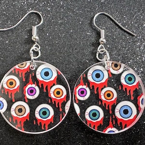 Puede incluir: Un par de pendientes redondos de acrílico transparente con fondo negro y un patrón de ojos coloridos con pintura roja goteando. Los pendientes tienen ganchos plateados.
