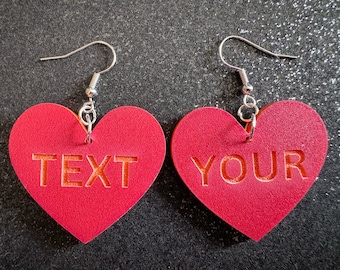 May include: A pair of red heart-shaped earrings with silver hooks. The left earring says "TEXT" and the right earring says "YOUR".