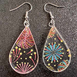 Puede incluir: Un par de pendientes de plata en forma de lágrima con fondo negro y diseños de fuegos artificiales coloridos. Los pendientes tienen un acabado transparente y brillante.