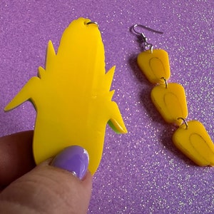 May include: A pair of yellow dangle earrings with a corncob design. The earrings are made of plastic and have a green outline.