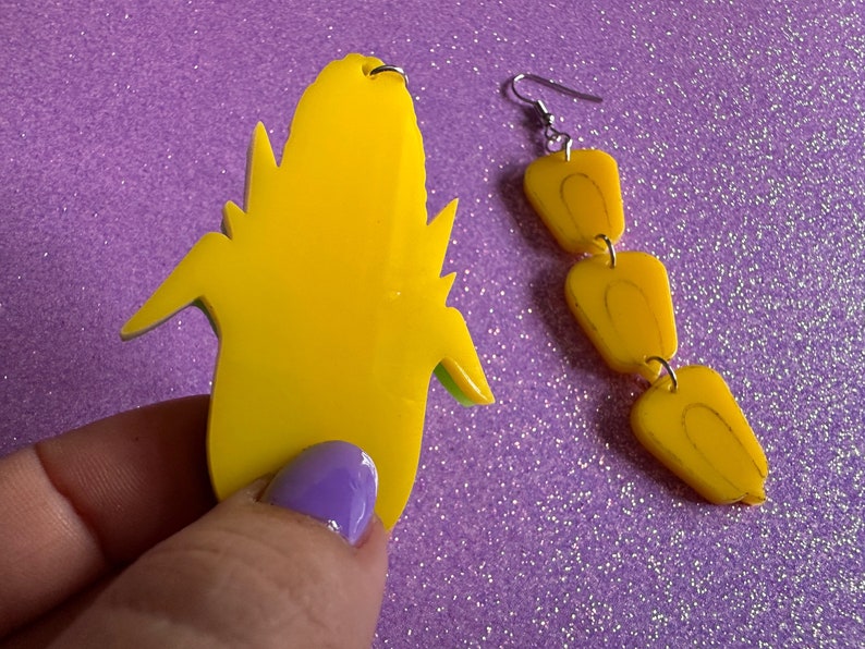 May include: A pair of yellow dangle earrings with a corncob design. The earrings are made of plastic and have a green outline.