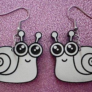 May include: A pair of white snail earrings with black outlines and large black eyes. The snails have a small smile and are attached to silver hooks.