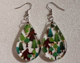 May include: A pair of teardrop-shaped earrings with a clear background and a pattern of green trees and brown Bigfoot figures.