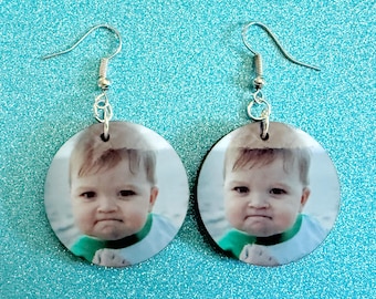 May include: A pair of round earrings featuring a baby's face with a slightly grumpy expression. The earrings have silver-colored hooks and are set against a sparkly, light blue background.