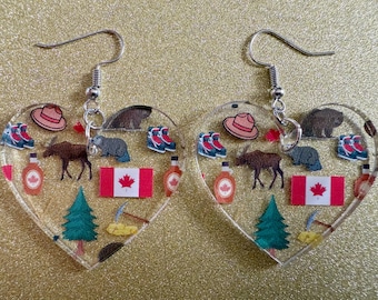 May include: Heart-shaped earrings with a clear background, featuring Canadian-themed icons. The earrings showcase a red and white Canadian flag, a moose, a beaver, a hat, maple syrup bottles, and other symbols.