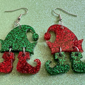 May include: A pair of elf-themed dangle earrings. One earring features a green glitter elf hat with red glitter elf shoes. The other earring features a red glitter elf hat with green glitter elf shoes.