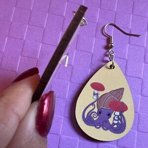 May include: A pair of wooden earrings shaped like teardrops. The earrings are painted with a light brown background and feature a purple octopus with a brown snail shell on its head. The octopus is holding two red mushrooms with white stems. The earrings have silver hooks.
