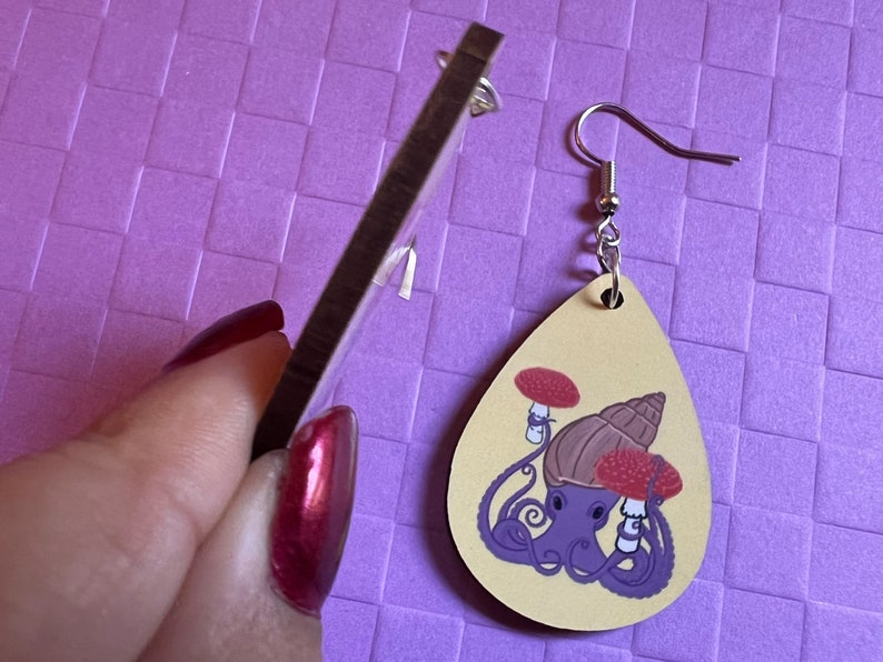 May include: A pair of wooden earrings shaped like teardrops. The earrings are painted with a light brown background and feature a purple octopus with a brown snail shell on its head. The octopus is holding two red mushrooms with white stems. The earrings have silver hooks.