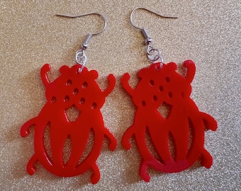 May include: A pair of red beetle-shaped earrings with silver hooks. The earrings are made of a shiny, smooth material and have a detailed design.