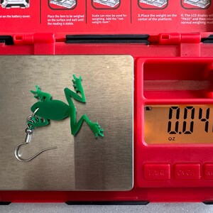 May include: A green frog earring with a silver hook is placed on a digital scale. The scale reads 0.042 ounces.