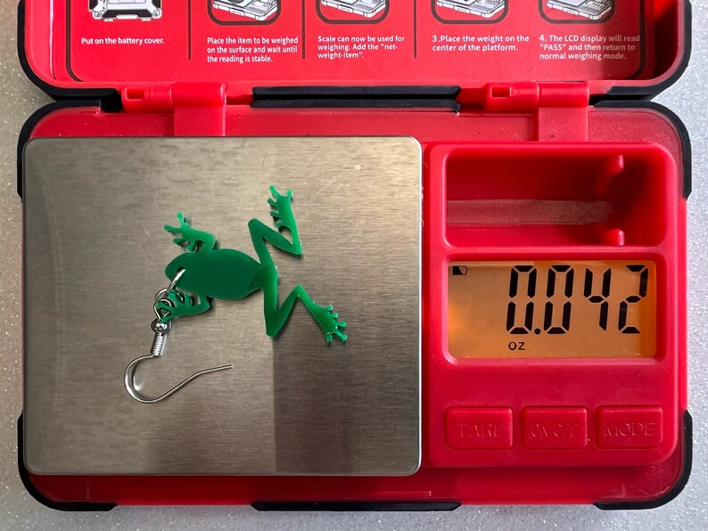 May include: A green frog earring with a silver hook is placed on a digital scale. The scale reads 0.042 ounces.