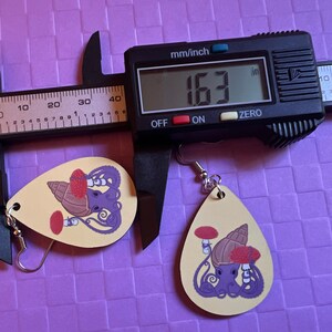 May include: Two teardrop shaped earrings with a yellow background. The earrings feature a purple octopus with a red mushroom and a brown seashell. The earrings are measured with a digital caliper that reads 163 inches. The earrings are on a purple background.