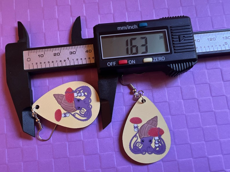 May include: Two teardrop shaped earrings with a yellow background. The earrings feature a purple octopus with a red mushroom and a brown seashell. The earrings are measured with a digital caliper that reads 163 inches. The earrings are on a purple background.