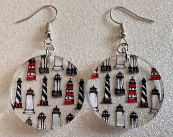 May include: A pair of round, clear acrylic earrings with a white background and a repeating pattern of black and red lighthouses. The earrings have silver hooks.