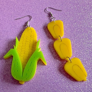 May include: A pair of earrings featuring a yellow corn cob with green husks and a yellow dangle earring with three sections.