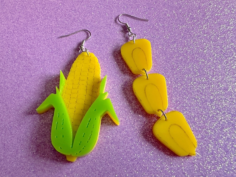 May include: A pair of earrings featuring a yellow corn cob with green husks and a yellow dangle earring with three sections.