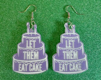 May include: A pair of purple cake-shaped earrings with white frosting and the text "Let them eat cake" on each earring.