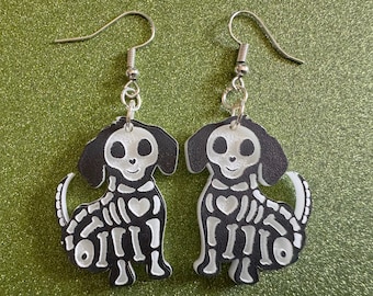 May include: A pair of dangle earrings featuring a black and white skeleton dog design. The dog is sitting with its tail curled up and has a heart on its chest. The earrings are made of silver metal.