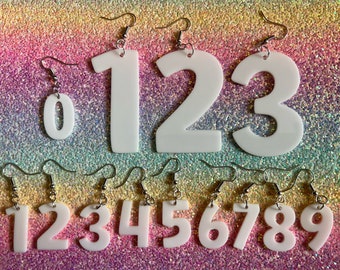 May include: White acrylic earrings shaped like the numbers 0 through 9. The earrings are on a glittery background.