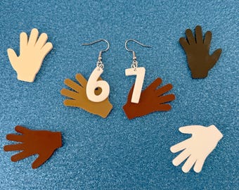 May include: Earrings featuring the numbers 6 and 7 in white, each attached to a hand-shaped charm. Additional hand-shaped charms in various skin tones are scattered around the earrings on a blue background. The earrings have silver-colored hooks.