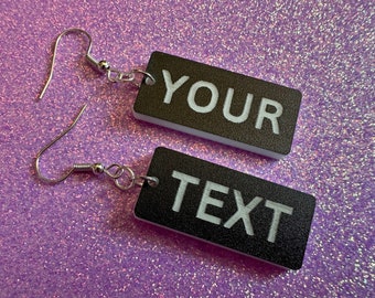 May include: A pair of black rectangular earrings with silver hooks. The earrings have white text that reads "YOUR TEXT".