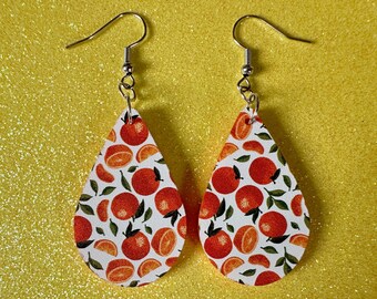 May include: A pair of teardrop-shaped earrings with a white background and a pattern of orange slices and leaves. The earrings are made of metal and have silver hooks.