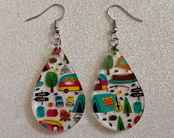 May include: A pair of teardrop-shaped earrings with a camping theme. The earrings are clear and feature a colorful pattern of camping equipment, including a tent, a camper, a campfire, and a guitar. The text "Camp Lake" is visible on the earrings.