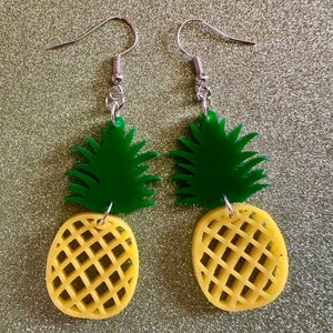 May include: A pair of pineapple earrings with green leaves and yellow pineapple slices. The earrings are made of acrylic and have silver hooks.