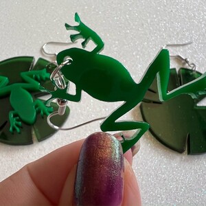 May include: A pair of green frog earrings with a lily pad design. The frogs are made of acrylic and have a shiny finish. The earrings are attached to silver hooks.