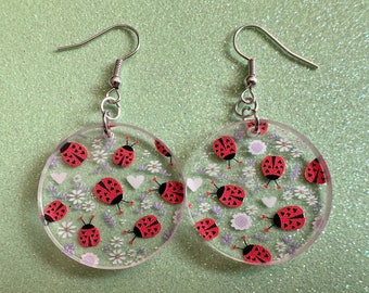 May include: A pair of round, clear resin earrings with a white and purple floral pattern and red ladybugs. The earrings have silver hooks.